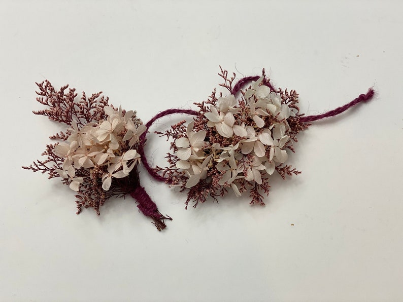 May include: Two small bouquets of dried flowers tied with burgundy twine. The bouquets are made of white hydrangeas and pink sprigs.
