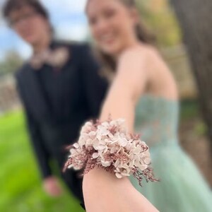 May include: A close-up of a wrist with a delicate floral bracelet made of dried flowers. The bracelet is made of white and pink flowers with brown stems. The flowers are arranged in a cluster and are tied together with a thin ribbon.