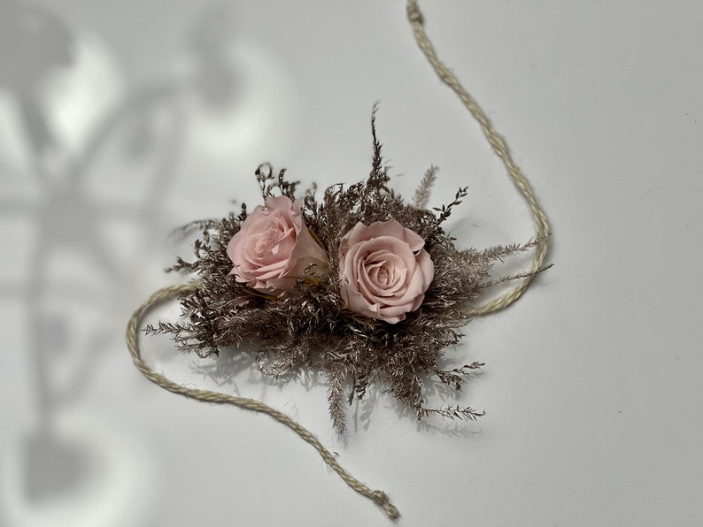 May include: Two pink roses are nestled in a bed of dried brown foliage, tied together with a natural twine string.