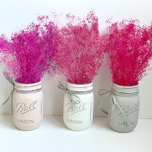 May include: Three mason jars painted in white and gray with a pink, light pink, and dark pink dried flower arrangement. The jars have the text "Ball Mason" on them.