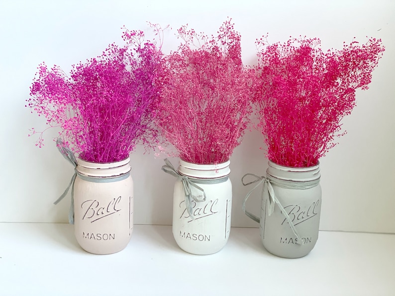 May include: Three mason jars painted in white and gray with a pink, light pink, and dark pink dried flower arrangement. The jars have the text "Ball Mason" on them.