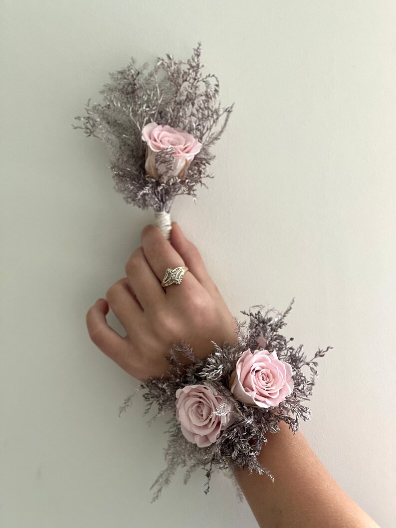 May include: A set of two floral wrist corsages with pink roses and gray dried foliage. The corsages are tied with white ribbon.