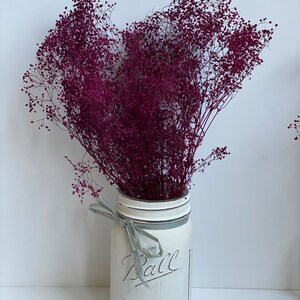 Peut inclure: Un bouquet de fleurs s&eacute;ch&eacute;es violettes dans un bocal en verre Mason peint en blanc avec un ruban gris autour du col. Le bocal porte l'inscription "Ball Mason" sur le devant.