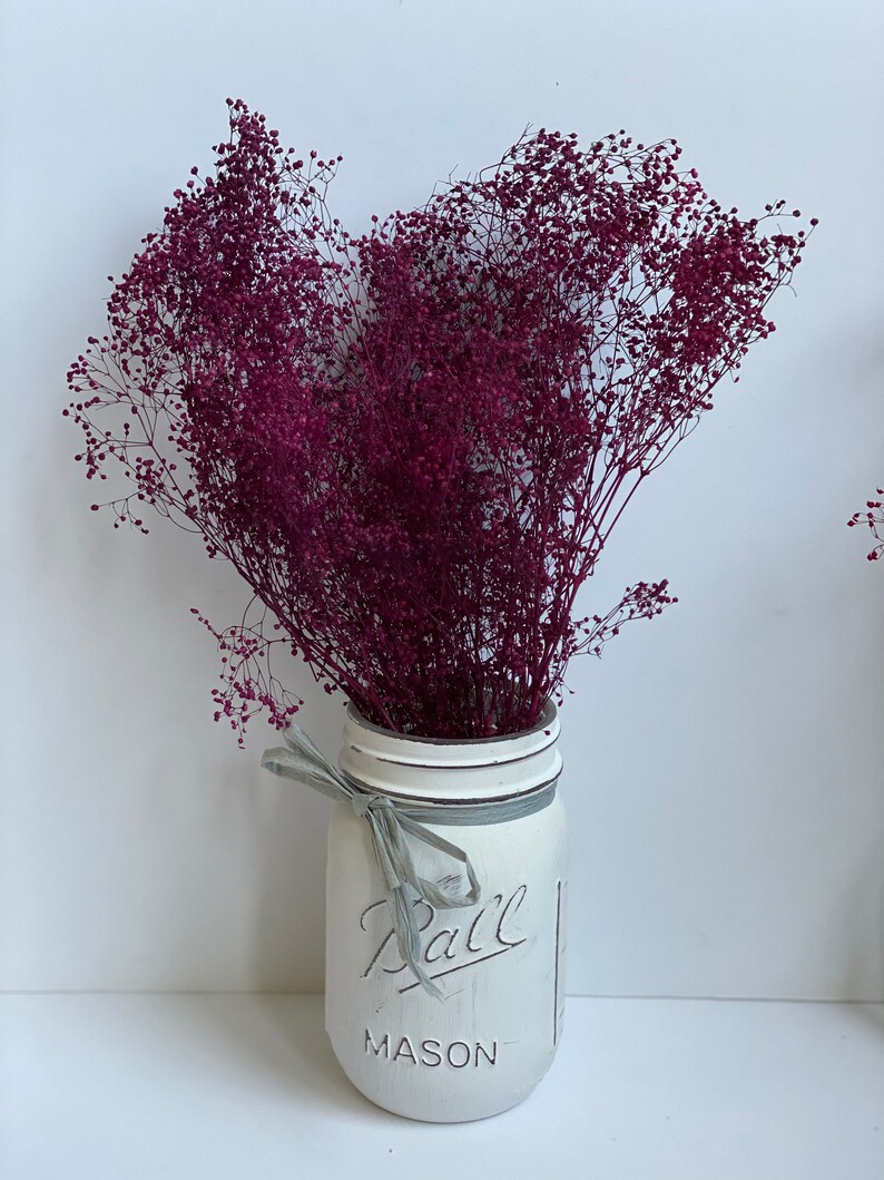 Peut inclure: Un bouquet de fleurs s&eacute;ch&eacute;es violettes dans un bocal en verre Mason peint en blanc avec un ruban gris autour du col. Le bocal porte l'inscription "Ball Mason" sur le devant.