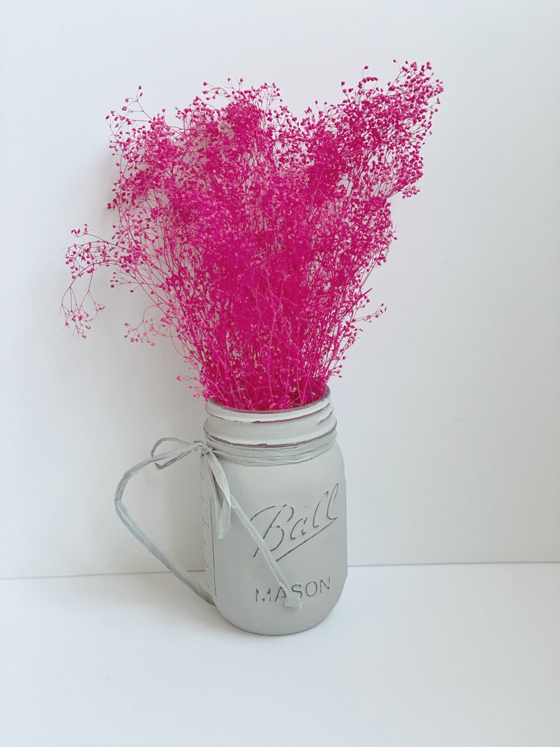May include: A gray painted mason jar with a white ribbon and the text "Ball Mason" on the front. The jar is filled with pink dried flowers.