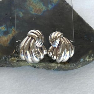 May include: A pair of shiny silver earrings. Each earring features a knotted design with a ribbed texture, reflecting light. The earrings are displayed on a dark, textured surface, showcasing their polished appearance.