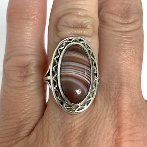 May include: A silver ring with an oval-shaped agate stone. The stone has brown, white, and gray stripes.