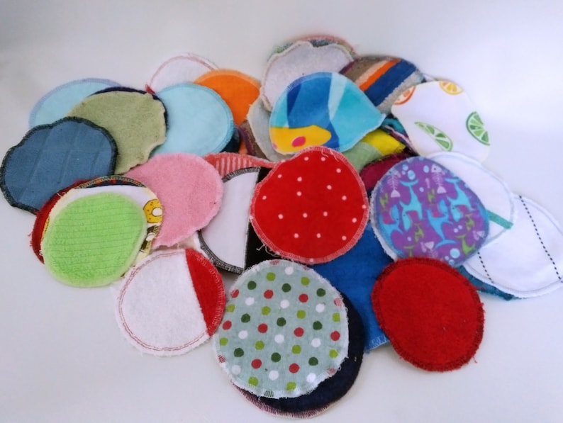 Op de afbeelding: Een verzameling kleurrijke, herbruikbare wattenschijfjes in diverse patronen en kleuren. De schijfjes bevatten effen kleuren zoals rood en blauw, en patronen met stippen, strepen en bloemmotieven. De randen zijn afgewerkt met stiksels.