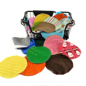 Op de afbeelding: Een verzameling kleurrijke, herbruikbare gezichtsreinigingspads in verschillende kleuren en patronen, waaronder geel, oranje, groen en rood. Sommige pads zijn effen, terwijl andere strepen of vakantie-thema's hebben. Ze worden in en rond een zwarte draadmand weergegeven.