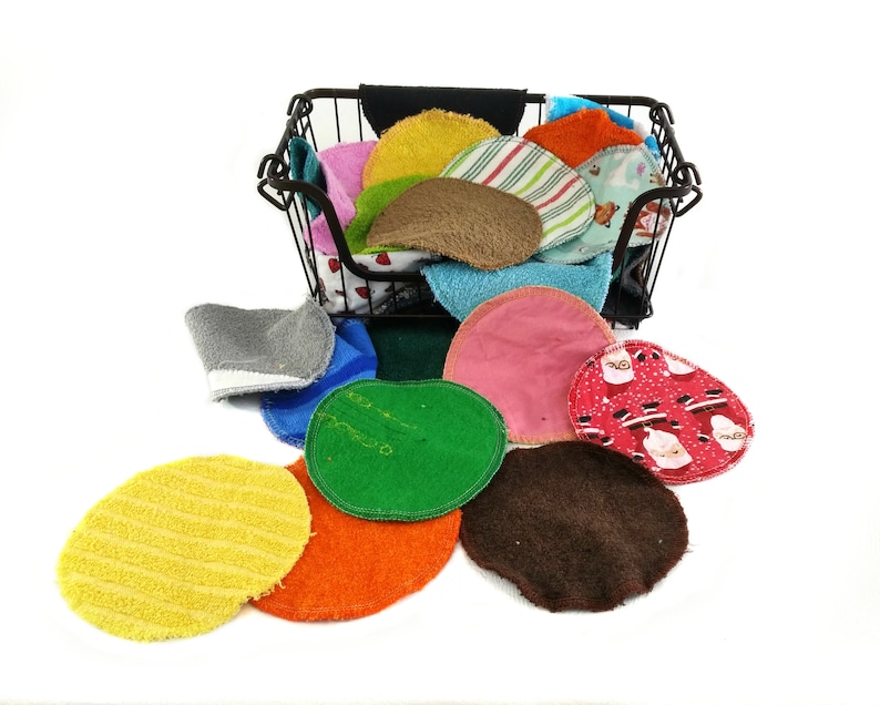 Op de afbeelding: Een verzameling kleurrijke, herbruikbare gezichtsreinigingspads in verschillende kleuren en patronen, waaronder geel, oranje, groen en rood. Sommige pads zijn effen, terwijl andere strepen of vakantie-thema's hebben. Ze worden in en rond een zwarte draadmand weergegeven.