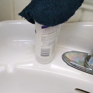 Op de afbeelding: Een witte fles met een etiket en een donkerblauwe washand die erop ligt. De fles staat in een witte wastafel met een chromen kraan.