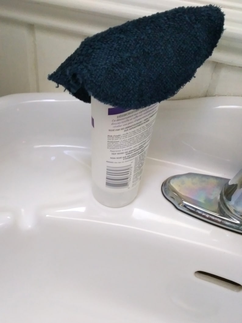 Op de afbeelding: Een witte fles met een etiket en een donkerblauwe washand die erop ligt. De fles staat in een witte wastafel met een chromen kraan.