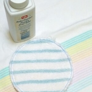 Op de afbeelding: Een witte plastic fles Life Baby Powder en een ronde borstvoedingspad met blauw-witte strepen. De borstvoedingspad is gemaakt van zacht materiaal en heeft een gestikte rand. De babypoederfles heeft een witte deksel.