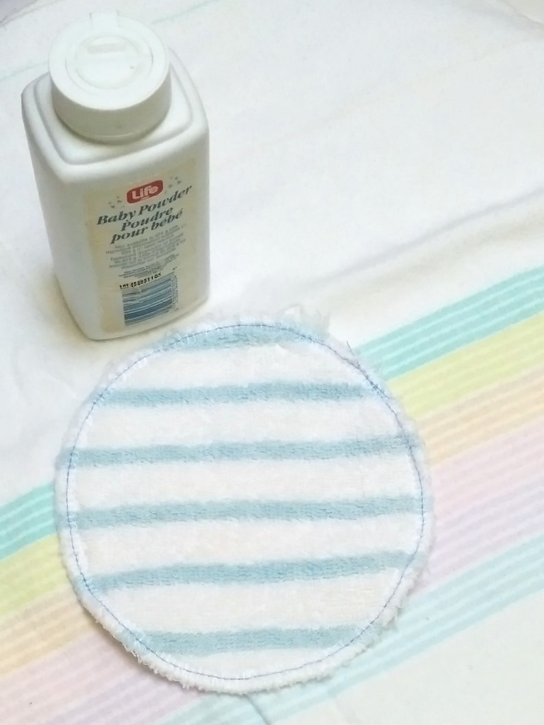 Op de afbeelding: Een witte plastic fles Life Baby Powder en een ronde borstvoedingspad met blauw-witte strepen. De borstvoedingspad is gemaakt van zacht materiaal en heeft een gestikte rand. De babypoederfles heeft een witte deksel.