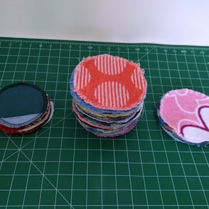 Op de afbeelding: Drie stapels herbruikbare gezichtsreinigingspads in verschillende kleuren en patronen. E&eacute;n stapel heeft een rood-wit stippenpatroon, een andere een roze bloemenpatroon en de derde een donkergroene stof. De pads liggen op een groene snijmat.
