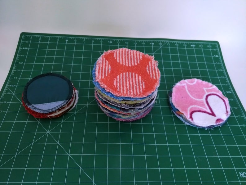Op de afbeelding: Drie stapels herbruikbare gezichtsreinigingspads in verschillende kleuren en patronen. E&eacute;n stapel heeft een rood-wit stippenpatroon, een andere een roze bloemenpatroon en de derde een donkergroene stof. De pads liggen op een groene snijmat.