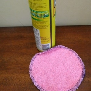 Op de afbeelding: Een ronde, roze, herbruikbare reinigingspad met een donkerpaarse gestikte rand ligt op een houten oppervlak. Een gele reinigingssprayfles staat op de achtergrond.