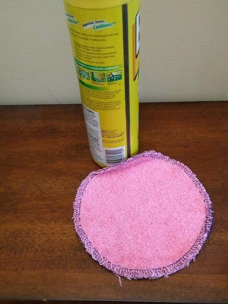 Op de afbeelding: Een ronde, roze, herbruikbare reinigingspad met een donkerpaarse gestikte rand ligt op een houten oppervlak. Een gele reinigingssprayfles staat op de achtergrond.