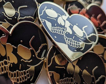 May include: A set of gold-colored heart-shaped pins with black enamel. Each pin features a pair of skulls with gold detailing, kissing each other.