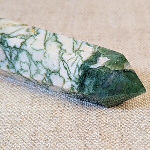 May include: A polished moss agate crystal tower featuring a pointed tip. The stone displays a white base color with intricate, branching dark green inclusions that resemble moss or fern patterns throughout the entire length of the mineral specimen.