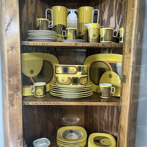 Puede incluir: Un armario de madera que muestra un juego de vajilla vintage amarillo y marrón. El juego incluye platos, tazas, tazas y fuentes para servir con diseños geométricos. El armario tiene puertas de vidrio y estantes de madera.