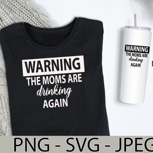 Op de afbeelding: Een zwart T-shirt met de tekst "WARNING THE MOMS ARE drinking AGAIN" in witte letters. De tekst "WARNING" staat in een roze rechthoek. De afbeelding toont ook een witte tumbler met dezelfde tekst in zwarte letters.