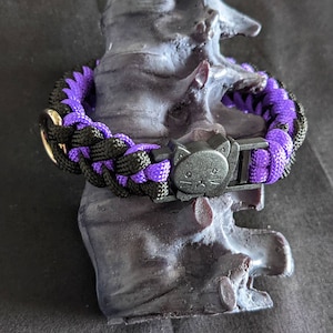Op de afbeelding: Een gevlochten halsband voor huisdieren in zwart en paars, met een zwarte katvormige gesp. De halsband wordt gepresenteerd op een donkere, getextureerde ondergrond. De halsband is ontworpen voor een klein huisdier.