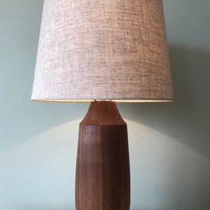 May include: A wooden table lamp with a dark brown base and a light gray fabric lampshade. The lamp has a tapered, cylindrical base and a wide, conical shade. The lamp is on a wooden surface against a light green wall.