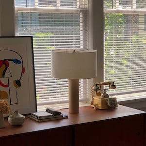 Peut inclure: Une lampe blanche avec une base cylindrique est posée sur une table en bois à côté d'un téléphone rotatif vintage. La lampe a un abat-jour blanc et est placée devant une fenêtre avec des stores blancs.