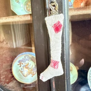 May include: A small, beige, woven fabric stocking ornament hanging from a metal cabinet key. The stocking features two red, square-shaped patches with visible white stitching on the upper leg and the heel area, and a ribbed cuff at the top.