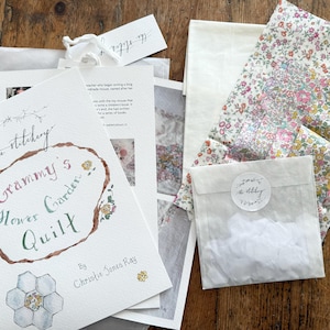May include: A craft kit titled Grammy's Flower Garden Quilt by Christie Jones Ray, featuring a printed instruction booklet, a piece of floral patterned fabric with small pink, yellow, and green blossoms, and a small bag of white fabric pieces.