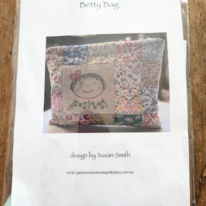 May include: A sewing pattern for a Betty Bag by Susan Smith, enclosed in a clear plastic sleeve. The paper insert features the text Betty Bag, design by Susan Smith, and an email address, alongside a photo of a patchwork bag with a central embroidered face.