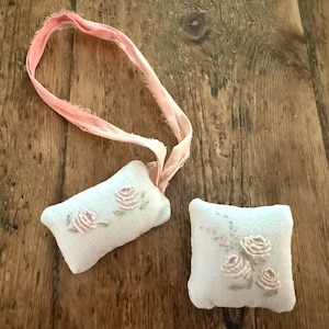 May include: Two small, rectangular, off-white fabric cushions featuring delicate embroidered pink rose motifs with green stems. One cushion is attached to a long, frayed, light pink ribbon loop, while the other is a standalone square-shaped piece.