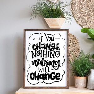 May include: A framed print with a white background and black text that reads "If you change nothing nothing will change". The print is hanging on a wall with a plant in a wooden planter and a woven wall hanging.