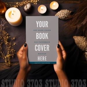 May include: A mockup of a 6x9 paperback book cover held by hands, with text that reads "YOUR BOOK COVER MOCKUP HERE". The cover is gray with a teal border. The background includes candles, dried plants, and decorative elements.