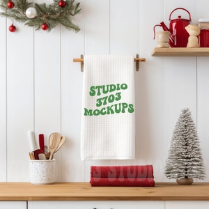 May include: White kitchen towel hanging on a wooden rack with the text "STUDIO 3703 MOCKUPS" in green letters. The towel is in a kitchen setting with a small Christmas tree, red and white ornaments, and a red teapot.