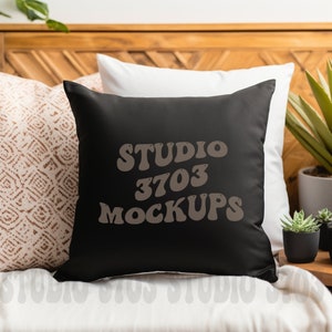 Puede incluir: Cojín cuadrado negro con el texto "STUDIO 3703 MOCKUPS" en marrón, con una fuente retro. El cojín está sobre una cama blanca con otras almohadas y plantas en el fondo.