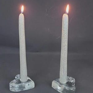 Può includere: Due candele argentate e sottili accese, in portacandele di vetro trasparente. Le candele hanno una consistenza glitterata. I portacandele sono a forma di cuore. Le fiamme sono di un arancione brillante su uno sfondo scuro. Le candele sono alte circa 20 cm.