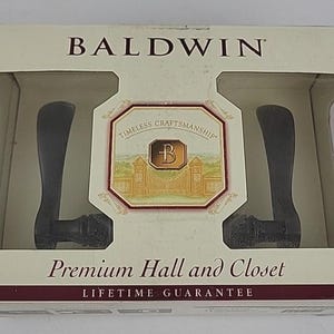 May include: A package of Baldwin door handles. The box is cream-colored with the brand name at the top. The handles are dark gray and are displayed in a clear plastic case. The box reads "Premium Hall and Closet" and "Lifetime Guarantee."