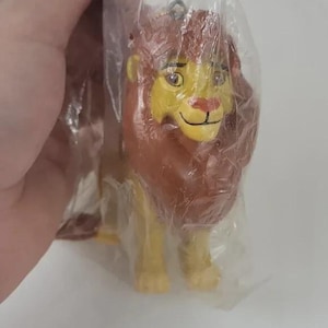 May include: A plastic figurine of a lion, featuring a brown mane, yellow face, and yellow legs. The lion is in a standing position and is inside a clear plastic bag. The figurine has a smiling expression.