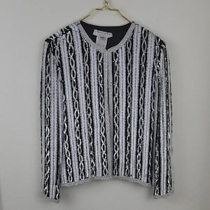 May include: A black and white sequined jacket with a vertical striped pattern. The jacket has long sleeves and a V-neckline. The design includes silver and black sequins arranged in a repeating pattern. The jacket is hanging on a gold hanger.