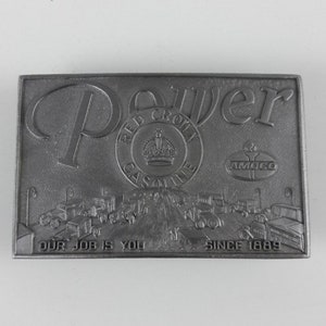 Puede incluir: Una hebilla de cinturón de plata con una imagen en relieve de una carretera con coches y el texto "Power Red Crown Gasoline Amoco Our Job Is You Since 1889".