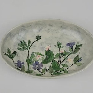 May include: Oval ceramic serving dish with a hand-painted floral design. The dish features a light blue background with green leaves, purple and pink flowers. The rim is a lighter shade of blue.