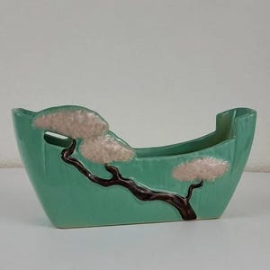 May include: A turquoise ceramic planter with a stylized tree design. The tree features a dark brown trunk and branches with three light pink, textured cloud-like shapes. The planter has a rectangular shape with curved sides.