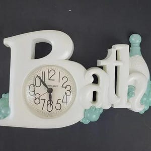 May include: White wall clock in the shape of the word "Bath". The "B" forms the clock face, with black numbers and hands. The clock is decorated with light blue bubbles and a bottle.