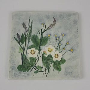 May include: Square ceramic tile featuring a botanical design. The artwork showcases white flowers with yellow centers, green leaves, and sprigs of purple and green foliage against a light blue background. The tile has a slightly textured appearance.
