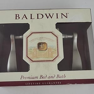 Puede incluir: Un juego de manijas de puerta Baldwin en su embalaje original. La caja es blanca con el nombre de la marca en letras grandes. El embalaje incluye las palabras "Timeless Craftsmanship" y "Premium Bed and Bath". Las manijas son plateadas.