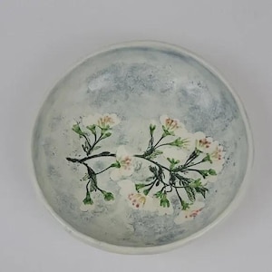 Puede incluir: Un cuenco pequeño y redondo de cerámica con un fondo moteado azul claro y blanco. El cuenco presenta un delicado diseño floral de flores blancas y hojas verdes. El borde del cuenco es blanco liso.