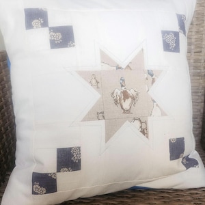 Puede incluir: Cojín cuadrado blanco con un diseño en forma de estrella en el centro, con una ilustración de pato. La estrella está rodeada por un fondo blanco y acentuada con cuadrados estampados florales azules. El cojín está sobre una silla de mimbre.