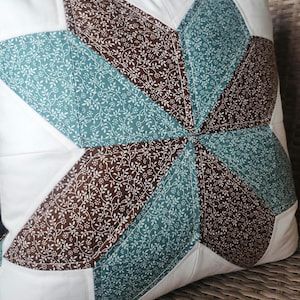 May include: Square cushion with a starburst design. The cushion has a white background with a central star pattern in teal and brown floral fabric. The cushion is placed on a woven surface.